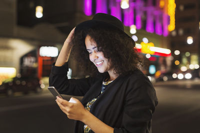 Smiling young woman reading text messaging on smart phone in city at night