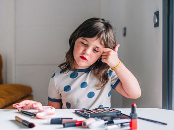 Child with red lips and closed eyes making up eyelid against various beauty products at home