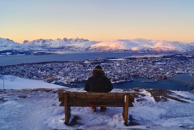 Rear view of man sitting on snowcapped mountain against sky in city