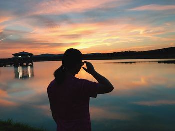 Silhouette woman standing by lake with orange clouds reflection at sunset