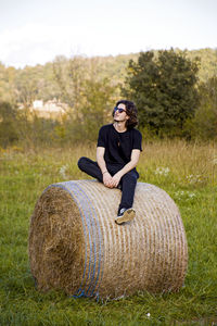 Full length of young man wearing sunglasses while sitting on hay bale against clear sky