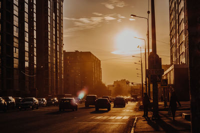 Cars on city street by buildings against sky during sunset