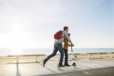 Father and son riding scooter on beach promenade at sunset