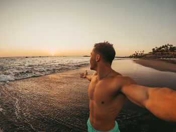 Shirtless man standing at beach against sky during sunset