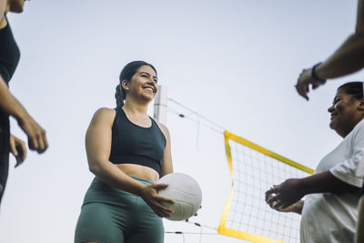 Low angle view of smiling woman playing volleyball with friends against sky