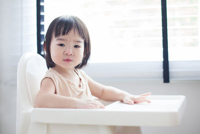 Portrait of cute baby girl sitting on high chair against window at home