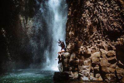 Side view of unrecognizable female traveler relaxing and enjoying fresh air while sitting on rough rocky slope near splashing waterfall in sunny day in los chorros municipal recreation park in costa rica