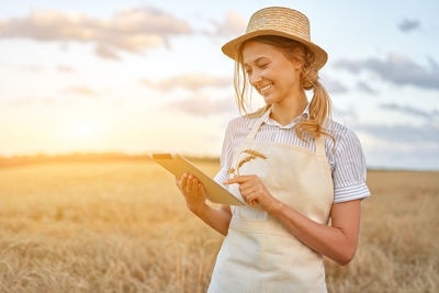 Smiling woman wearing hat using digital tablet while standing on field against sky