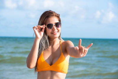 Young woman wearing sunglasses gesturing while standing against sea