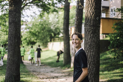 Side view portrait of smiling teenage boy standing by trees