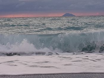 Waves rushing towards shore against sky during sunset