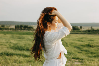 Beautiful young woman with hands in hair standing on land against sky