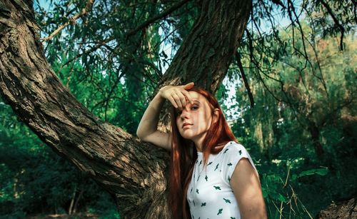 Portrait of young woman leaning on tree trunk while standing in forest