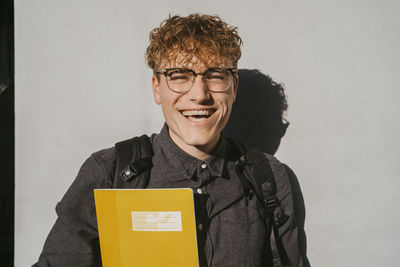 Portrait of happy blond male student wearing eyeglasses holding book against gray wall