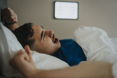 Teenage boy yawning on bed while waking up in morning at home