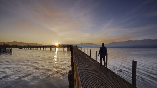 Man standing on pier over lake against sky during sunset