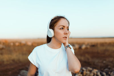 Young woman listening over headphones while sitting on land against sky