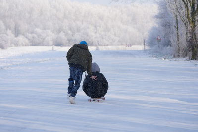 Rear view full length of man pushing person on skateboard at snow covered field