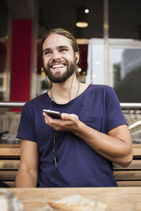 Smiling man listening music through smart phone while sitting at sidewalk cafe