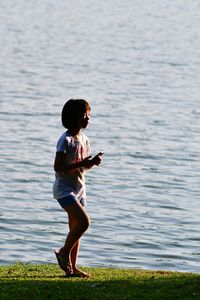 Side view of girl holding mobile phone while standing by lake