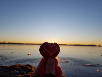 Hand holding heart shape at beach against sky during sunset