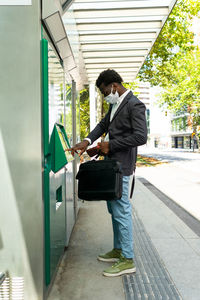 Side view of african american businessman in protective mask with suitcase using street atm terminal in barcelona city