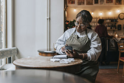 Mature female owner using mobile phone while sitting with diary at table in cafe