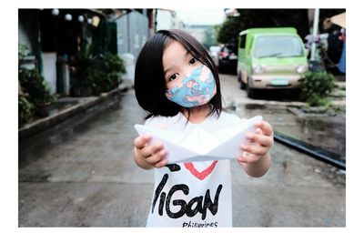Portrait of girl wearing mask holding paper boat while standing outdoors