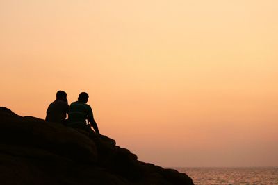 Rear view of men on rock by sea against sky during sunset