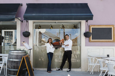 Smiling restaurant owners with arms crossed standing by glass window