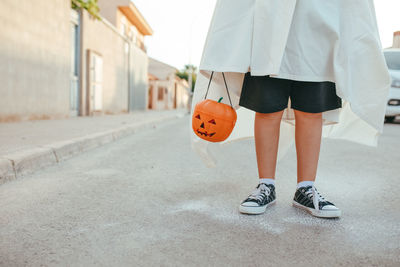 Cropped unrecognizable kid in white ghost costume and with plastic jack o lantern standing in city on halloween in autumn