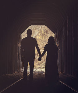 Rear view of silhouette couple holding hands while standing on railroad track in tunnel