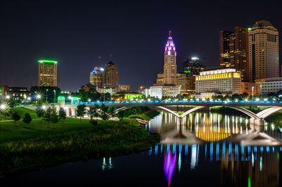 Illuminated bridge over river by buildings against sky at night