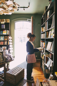 Side view of woman reading book while standing near bookshelf at store