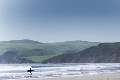 Surfer walking at beach by green mountains against clear blue sky