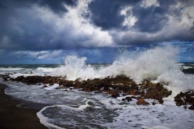 Waves splashing on rocks at beach against cloudy sky