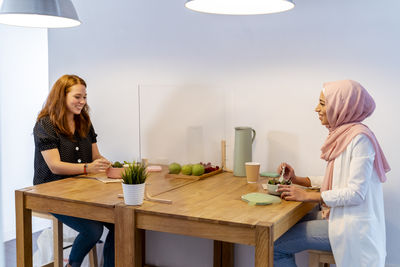 Smiling women having food while sitting at screen partition table in office