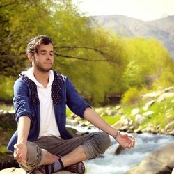 Man meditating on rock by flowing stream in forest
