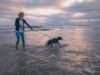 Woman with dog wading in sea against sky during sunset