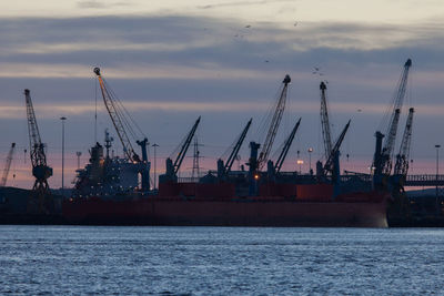 Cranes at commercial dock against cloudy sky at dusk