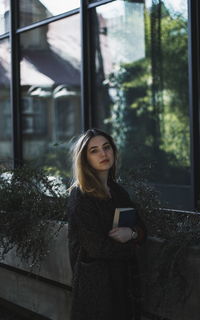 Young woman holding book while standing by window