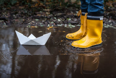 Low section of boy standing by paper boat in puddle