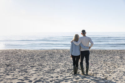 Man with arm around woman looking at sea standing on sand