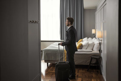 Side view of businessman standing with luggage by bed in hotel room