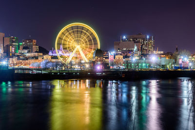 Illuminated ferris wheel by river against buildings at night