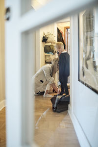 Side view of woman kneeling on floor and assisting son in wearing shoe seen through door