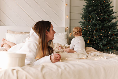 Moody sad little daughter child offended by mom sitting on the bed at the christmas tree at home