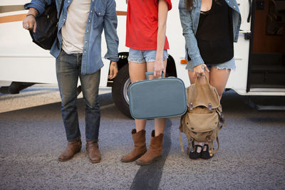 Low section of friends carrying luggage while standing on road against camper van