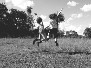 Rear view of siblings jumping on grassy field against cloudy sky