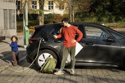 Father with hand on hip charging electric car while daughter playing at station